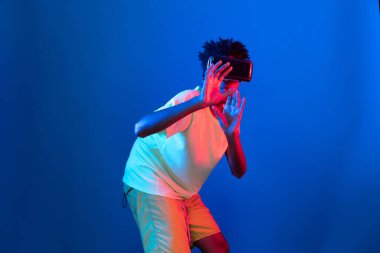 Genç Afrikalı Amerikalı basketbol oyuncusu, metaevren dünyasında dijital fütüristik teknolojiyi birbirine bağlayan mavi neon ışıkta zıplayan VR pozu takıyor. Entrika.