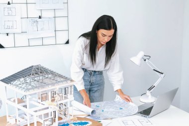 Genç, beyaz, güzel bir mimarın portresi toplantı masasına yerleştirilen ev modeli, plan ve dizüstü bilgisayarın yapısını karşılaştırmaya odaklanıyor. Şah, teftiş, tahmin konsepti. Tertemiz..