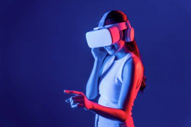 Siberpunk neon ışıklı cihazlarla çevrili zeki bir kadın metaevrene bağlı VR kulaklık takıyor, gelecekçi siber uzay teknolojisi. Sanal objeyi işaret eden bir kadın. Halüsinasyon.