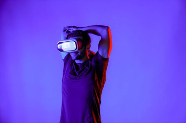 VR gözlüklü mutlu adam neon ışıklı arka planda egzersiz yapıyor ve kolunu esnetiyor. Günlük kıyafetleri olan beyaz bir insan sanal gerçeklik simülatörü teknolojisi kullanarak rahatlıyor ve egzersiz yapıyor. Sapma.