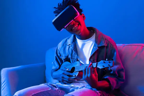 stock image Young African American singer using through VR glasses enjoy singing song with guitar playing on meta blue neon light wall virtual reality metaverse fantasy technology sitting on sofa. Contrivance.