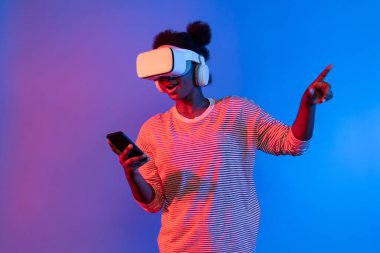 VR kulaklıklı ve akıllı telefonlu genç Afrikalı kadın canlı neon ışıklandırma arka planı sanal gerçekliği ve mobil bağlantı öğrenme teknolojisiyle metaevrene doğru yol alıyor. Entrika.