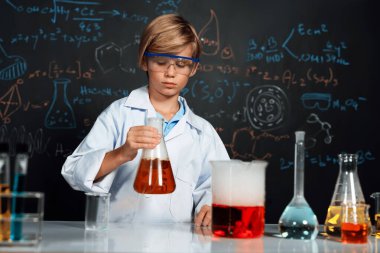 Laboratuvardaki sarışın çocuk laboratuvar önlüğü giyer ve STEM sınıfında kimya bilimi öğrenir. Öğrenci portakal suyu ile deney kabını tutuyor. Masaya bir sürü şişe koy. Etkinlik.