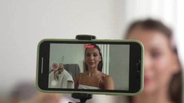 Genç sosyal medyum kadın moda videosu yapıyor. Blogcu kameraya selfie çubuğu ve ışık halkası kullanarak gülümserken, aynı zamanda ikna edici çevrimiçi kıyafetlerin izleyicilere veya takipçilere vlog satmasını sağlıyor. Düzenle