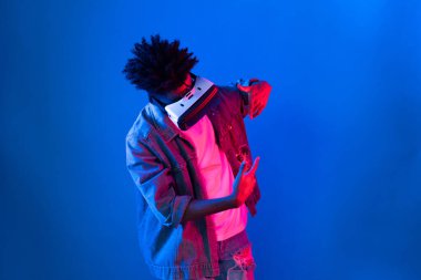 VR kullanan genç Afro-Amerikalı, boş uzay ürünlerini işaret ederek metaevren dünyasındaki mavi, neon ışıklı duvar fütüristik teknolojisi hakkında detaylı bilgi veriyor. Entrika.