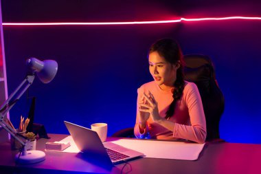 Geceleri sosyal medyada canlı yayında pozitif podcastla konuşan gülümseyen genç, güzel Asyalı etkileyici pembe neon ışıklı modern stüdyoda sunum için dizüstü bilgisayar kullanıyor. Taktik.