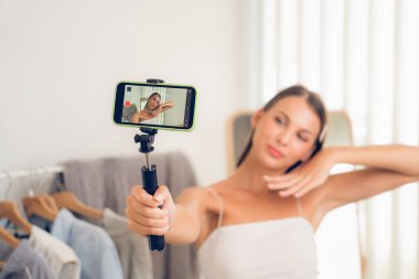 Sosyal medya için moda videosu hazırlayan genç bir kadın. Blogcu kameraya selfie çubuğu ve ışık halkası kullanarak gülümserken, aynı zamanda ikna edici online kıyafetlerin izleyicilere veya takipçilere satılmasını sağlıyor. Açılıyor