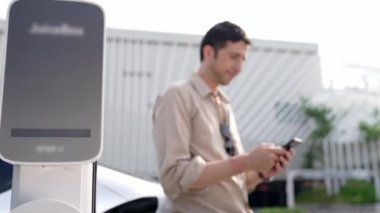 Ağır çekimde çalışan adam, alışveriş merkezinin otoparkındaki kamu EV şarj istasyonunda elektrik için akıllı telefon kullanıyor. EV aracıyla modern çevre ve sürdürülebilir kentsel yaşam tarzı
