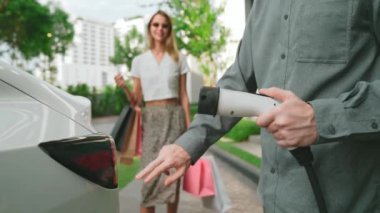 Yaz mevsiminde yeşil sürdürülebilir şehir bahçesinde ağır çekimde çalışan EV elektrikli araba yeşil sürdürülebilir şehirde yeniden şarj edilebilir elektrikli araç iç organları için alışveriş yapan genç çiftlerin yaşam tarzını gösteriyor.