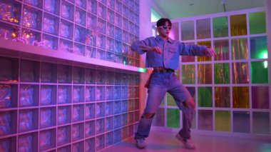 Gece kulübünde kameraya bakarken güneş gözlüklü Asyalı dansçı serbest dans ediyor. Mutlu sokak dansçısı dans ediyor ve renkli parlak neon ışığı ya da yol gösteren ışıkla ayak seslerini gösteriyor. Mutluluk.