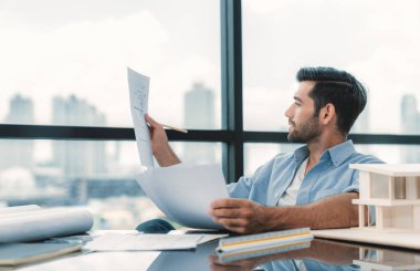 Modern ofisteki masanın üzerinde, yetenekli beyaz mimar mühendisi planları tutuyor. Tasarım ve ev modeli arasında karşılaştırıldığında akıllı bir iç mimar. Proje, plan, inşaat, proje. İzleme.
