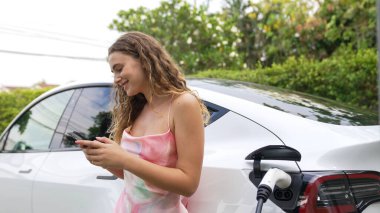 Genç bir kadın doğadaki EV şarj istasyonunda elektrik için akıllı telefon kullanıyor. Modern çevre ve EV araçlı sürdürülebilir otomobil taşıma yaşam tarzı. Eşzamanlama