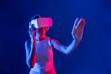 Çevresi siberpunk neon ışıklarıyla çevrili zeki bir kadın metaevrene bağlı VR kulaklık takıyor, gelecekçi siber uzay topluluğu teknolojisi. Sanal objeye el ile dokunan kadın. Halüsinasyon.