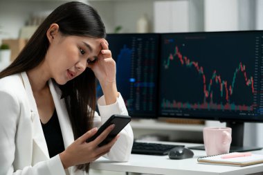 Stresli, Asyalı genç ve güzel iş kadını akıllı telefona odaklanıyor, modern ofisteki bilgisayar ekranında dinamik borsa grafiğinde kar veya zarar etme riskini analiz ediyor. Taktik.