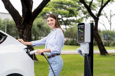 Açık yeşil şehir parkı manzaralı EV şarj istasyonundan genç kadın EV elektrikli araçları şarj ediyor. Çevre dostu şehir ulaşımı ve çevre dostu EV araba seyahati. Yükselt