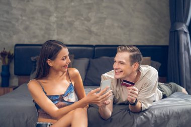 Genç çift, kredi kartıyla ödemek için internet üzerinden ödeme uygulaması ve dijital cüzdan kullanarak yatak odasında oturuyor. Mobil internet üzerinden alışveriş ve modern alımlar. Açılıyor