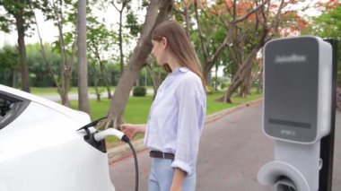 Yavaş çekimde genç bir kadın sonbaharda ulusal parktaki EV şarj istasyonunda EV elektrikli arabaları şarj ediyor. Tatil ve sonbahar sezonunda çevre dostu arabalarla seyahat. Yararlı