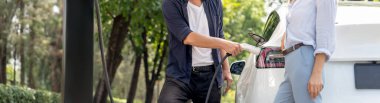 Odaklanmış EV arabanın pili, tatilde ulusal park ve yeşillik ormanlarında çevre dostu elektrikli arabalarla seyahat eden sevgili çiftin bulanık arka planına şarj oluyor. Yükselt