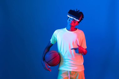 Genç Afrikalı Amerikalı basketbol oyuncusu, metaevren dünyasında dijital fütüristik teknolojiyi birbirine bağlayan mavi neon ışıkta zıplayan VR pozu takıyor. Entrika.