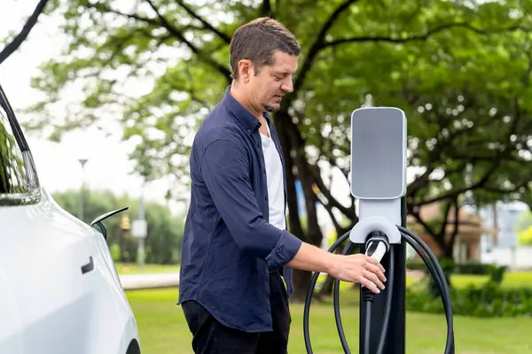 Açık yeşil şehir parkı manzaralı EV şarj istasyonundan EV elektrikli araçları şarj eden adam. Çevre dostu şehir ulaşımı ve çevre dostu EV araba seyahati. Yükselt