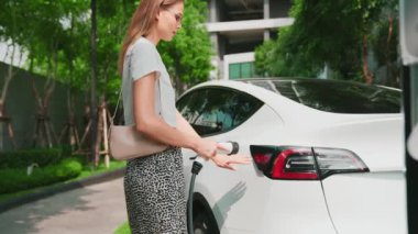 Şehir merkezindeki EV şarj istasyonunda elektrikli araba aküsünü yavaş çekimde şarj eden kadın elektrikli aracın iç organları için sürdürülebilir yeşil ve yenilenebilir temiz enerji yaşam tarzına sahip.