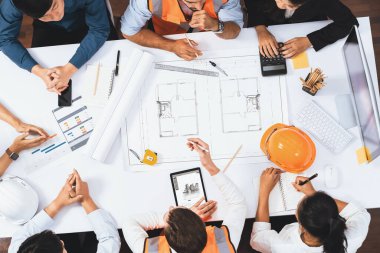Mimari proje üzerinde birlikte çalışan, inşaat planını gözden geçiren ve toplantı masasında plan oluşturan çeşitli inşaat mühendisi ve müşteri gruplarından oluşan üst düzey bir pankart. Tedbirli