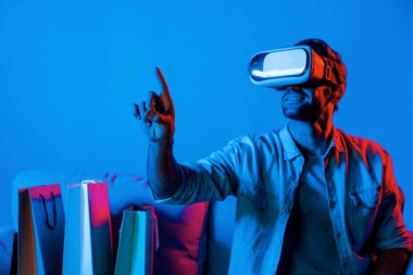 Video kameralı beyaz bir adam kanepeye oturmuş alışveriş torbalarıyla işaret ediyor. VR teknolojisi ve evde sanal gözlük kullanarak online alışveriş yapan kişi. Sapma.