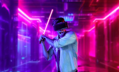 Lazer kılıcı tutan ve VR gözlük takan oyuncu. Beyaz adam, neon şatonun etrafında dikilirken görsel gerçeklik kulaklığı kullanıyor. Yenilik teknolojisi. Sapma.