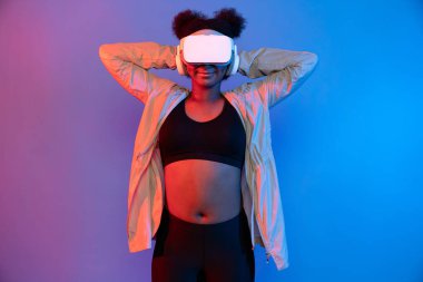 VR giyen genç Afrikalı spor kadın, dijital fütüristik teknolojiyi sanal gerçekliğe bağlayan parlak mavi neon ışıkları üzerinde poz veriyor. Entrika.