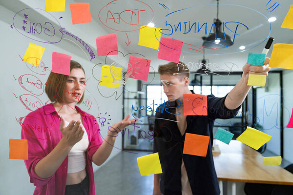 Couple of businesspeople discussing and brainstorming about solving marketing problem in front of glass board at business meeting by using mind map and sticky notes. Working together. Immaculate.