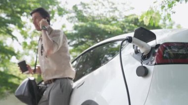 Yavaş çekim adamı ve EV elektrikli araba ile sürdürülebilir şehir ulaşımı ilkbahar bahçesindeki açık kahve kafeteryasında şarj oluyor, yeşil şehir sürdürülebilirliği ve çevre dostu EV aracı. Yararlı