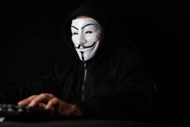 Kameraya bakan beyaz sırıtan isimsiz suçlu maskesi ana veritabanı sunucu sistemi aracılığıyla bilgisayar fidye şirketinin gizliliğinde başarıyla hacklenen virüs şifrelemesi yüklüyor. Tahmin et..