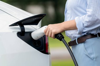 Açık yeşil şehir parkı manzaralı EV şarj istasyonundan genç kadın EV elektrikli araçları şarj ediyor. Çevre dostu şehir ulaşımı ve çevre dostu EV araba seyahati. Yükselt