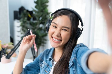 Sunucu kanalı genç ve güzel Asyalı kız akıllı telefonuyla selfie çekiyor. Modern stüdyo ofis konseptinde kulaklık takıyor. Sahne arkası yayıncısı sosyal medyada canlı yayın yapıyor. Taktik.