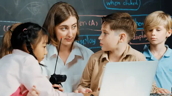 stock image Caucasian boy using laptop programing engineering code and writing program while young caucasian teacher holding controller in STEM technology classroom at blackboard written with prompt. Erudition.