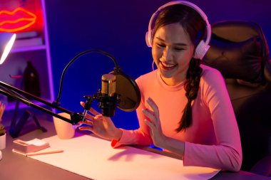 Smiling creative young beautiful Asian influencer talking in channel with positive podcast speech at night time, using laptop for presentation online at pink neon light modern studio. Stratagem. clipart