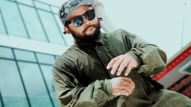 Asyalı hippi gökyüzü dövüşçüsüne dans ederken kameraya bakıyor. Çekici yakışıklı hippi ellerini alçak açılı kamerayla modern alışveriş merkezinde hip hop müziğine yöneltiyor. Açık hava sporu 2024. Hiphop.