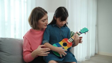 Kafkasyalı bir anne evde akustik müzik öğretirken ukulele çalan mutlu kız. Sevimli çocuk enstrüman öğreniyor. Kafkasyalı mutlu anne ve kız birlikte vakit geçiriyorlar. Pedagoji.