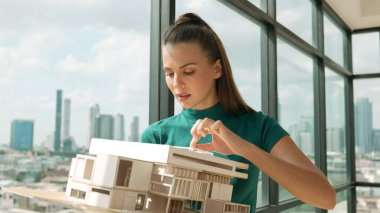 Genç akıllı mimar mühendisi ev modelini incelerken mimari modeli elinde tutuyor. Şehir manzaralı pencerenin yanında dururken profesyonel iç mimar ev inşaatını kontrol ediyor. İzleme