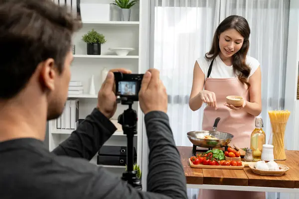 Stock image Cameraman recording to woman in chef influencer host cooking spaghetti with meat topped tomato sauce surrounded ingredients recipe, presenting special dish healthy food at modern studio. Postulate.