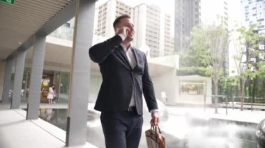 Modern koridorda yürürken telefonla konuşan mutlu proje yöneticisi. Parkta ya da yeşil şehirde yürürken finansal stratejiyi planlamak için pazarlama ekibini arayarak telefonu kullanıyor. Urbane.