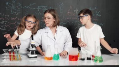 Öğretmen tavsiye verirken beyaz çocuk kimyasal sıvıyı karıştırıyor. Profesyonel eğitmen labaratuar kıyafeti giymiş, renkli solüsyonla dolu deney kabıyla sofrada çeşitli öğrencileri arıyor. Etkinlik.