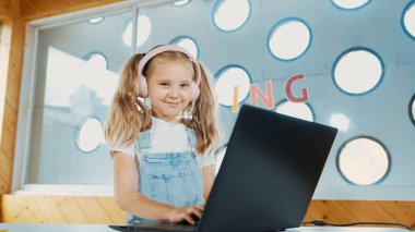 Little young cute girl using laptop to working or playing games. Student working on computer while looking and smiling at camera. Caucasian child attend online class by using headphone. Erudition. clipart