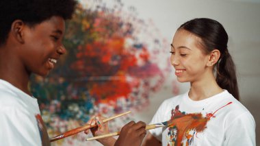 Resim dersinde, renkli duvarda, boyalı beyaz gömlekleri boyayarak gülümseyen farklı çocuklar. Tecrübe alışverişini temsil etmek, birbirimizi öğrenmek. Öğretim.