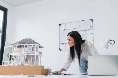 Genç, beyaz, güzel bir mimarın portresi toplantı masasına yerleştirilen ev modeli, plan ve dizüstü bilgisayarın yapısını karşılaştırmaya odaklanıyor. Şah, teftiş, tahmin konsepti. Tertemiz..