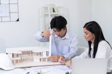 Profesyonel Asyalı erkek mimar ev modelinin uzunluğunu ölçmek için cetvel kullanırken, beyaz tenli genç meslektaşı ev modeliyle toplantı masasındaki verileri analiz etmek için dizüstü bilgisayar kullanıyor. Tertemiz..