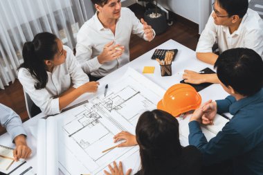 Mimari proje üzerinde birlikte çalışan çeşitli inşaat mühendisi ve müvekkil grupları, inşaat planını gözden geçirip toplantı masasında plan oluşturuyorlar. Tedbirli