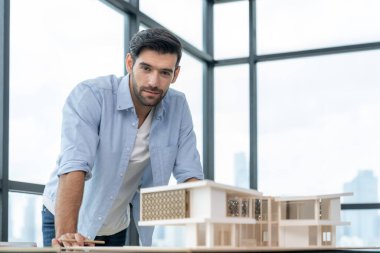 Ev modeliyle dikilirken kameraya bakan gülümseyen beyaz mimar mühendisi. Kalem tutarken ayakta duran zeki bir iç mimar. Proje planı, inşaat mühendisliği, işçi. İzleme.