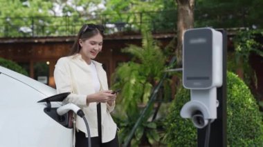 İlkbahar bahçesindeki açık kafede çalışan EV elektrikli araba şarjı, yeşil şehir sürdürülebilirliği ve çevre dostu EV otomobili için genç bir kadınla sürdürülebilir şehir seyahati elektrik ödüyor. Yararlı