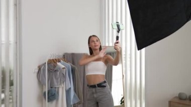 Genç sosyal medyum kadın moda videosu yapıyor. Blogcu kameraya selfie çubuğu ve ışık halkası kullanarak gülümserken, aynı zamanda ikna edici çevrimiçi kıyafetlerin izleyicilere veya takipçilere vlog satmasını sağlıyor. Düzenle